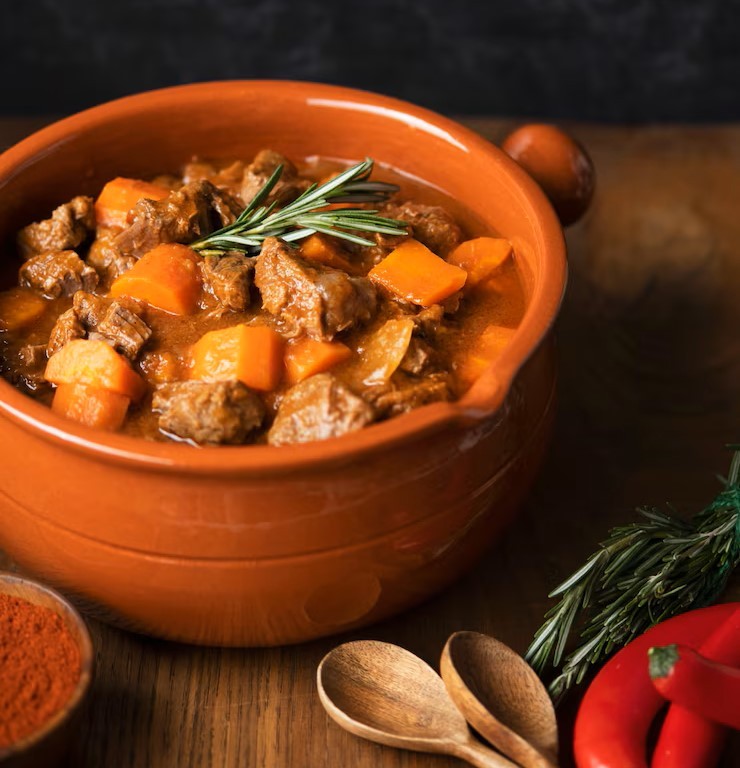 Warm beef Goulash (stew) in a deep orange pot next to red chilli, rosemary and two wooden spoons