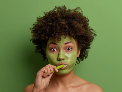 Young model wearing green face mask with toothbrush in her mouth, looking surprised