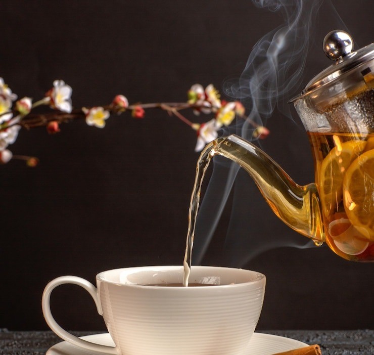 A cup of black tea being poured out of a glass tea pot with rising steam and background of flower buds