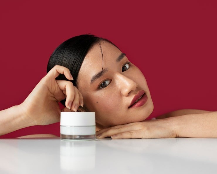 Young woman leaning on a white tabletop, touching a jar of moisturizer