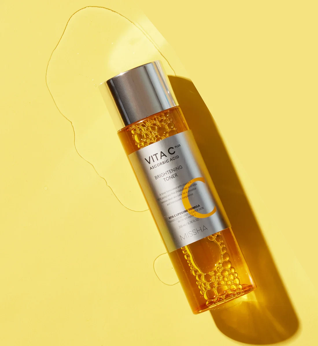 Missha Vita C Brightening Toner lying in a pool of toner on a yellow background