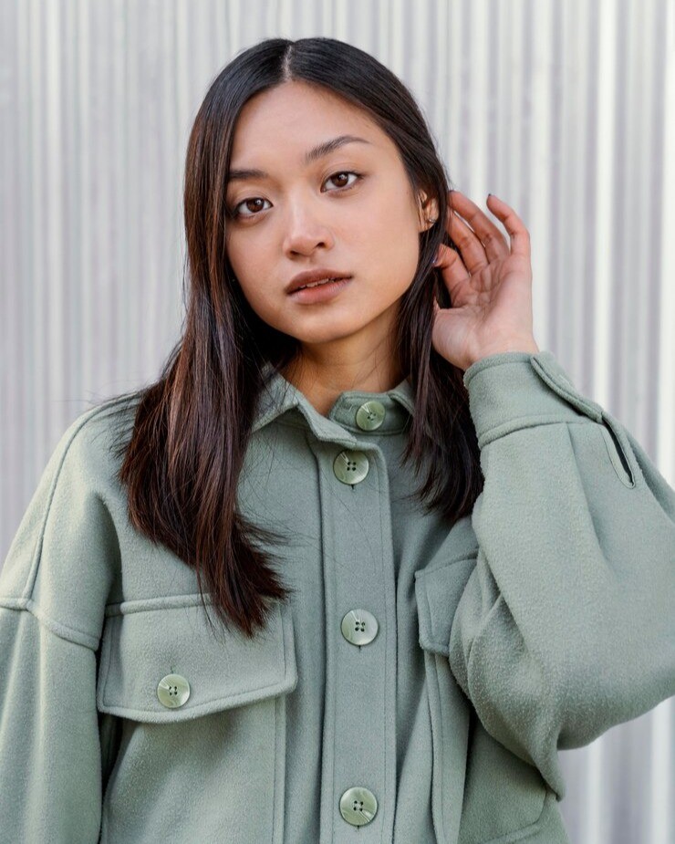 Young woman with black hair posing in front of steel background with green-blue trenchcoat