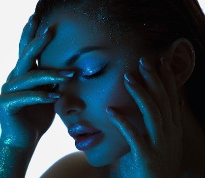 Model in dark-blue lighting covered in glitter and holding her face