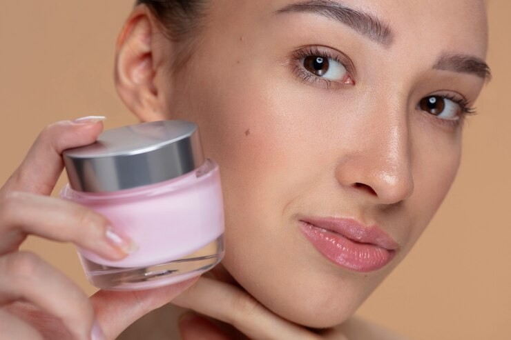 Young woman with brown eyes and light makeup holding a pink face cream jar to her cheek