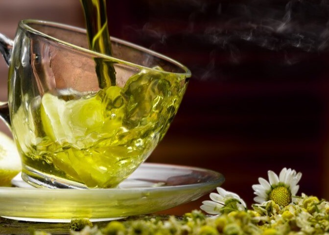 Pouring green tea into clear, glass mug with background of chamomile flowers.