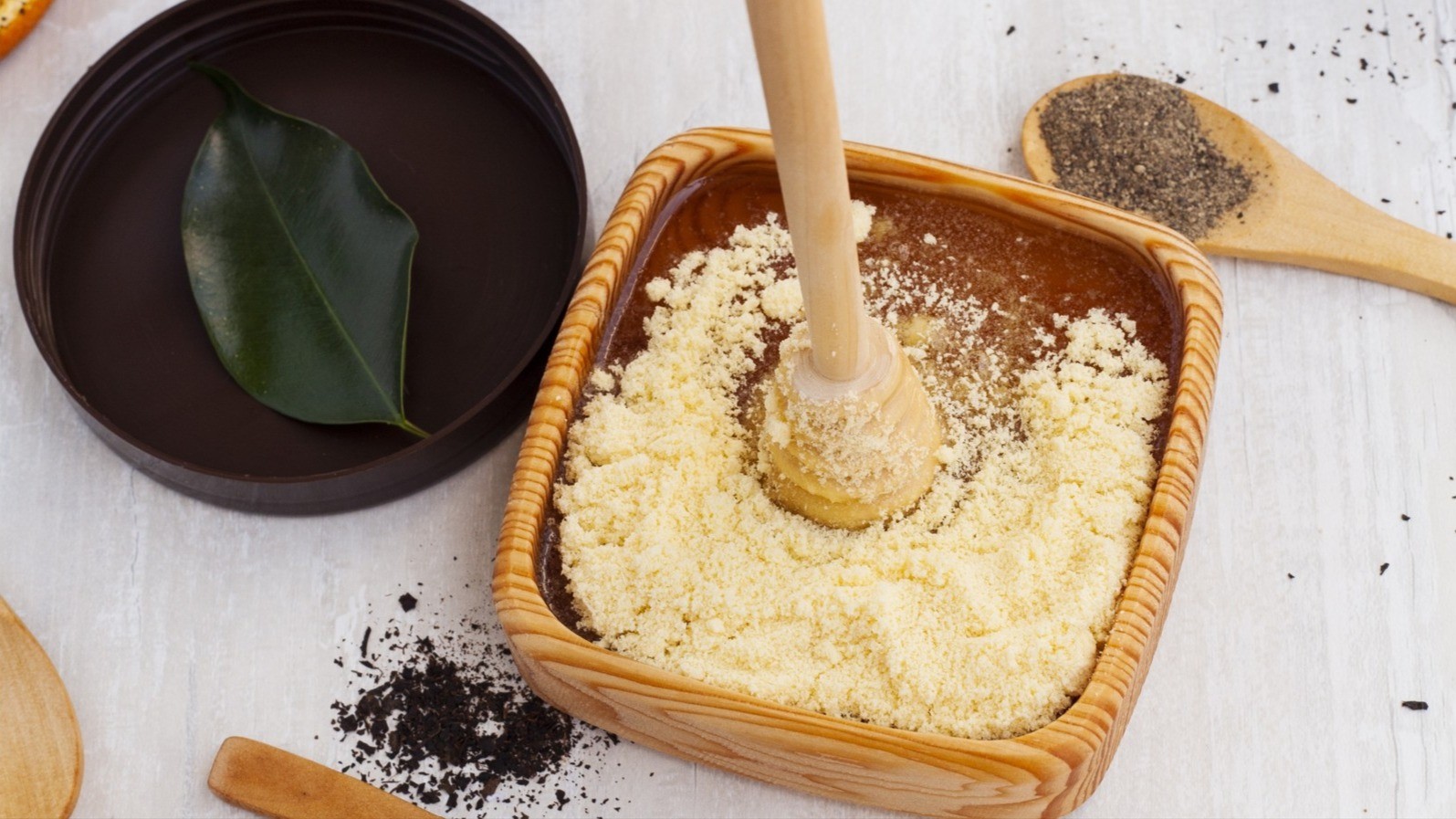 Exfoliating mixture of honey and flour being mixed with a wooden honey spoon, surrounded by various ingredients