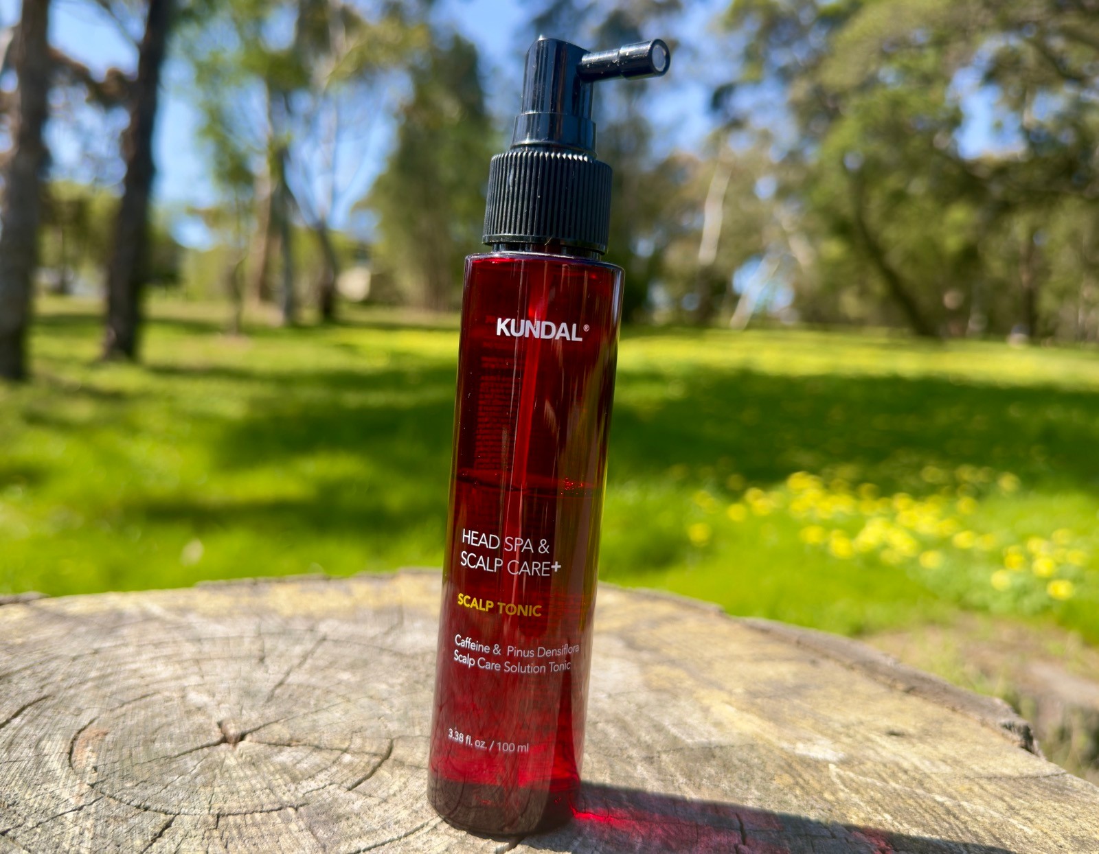 KUNDAL Head Spa & Scalp Care+ Scalp Tonic 100ml spray bottle standing upright on a wooden stump in an outdoor park setting