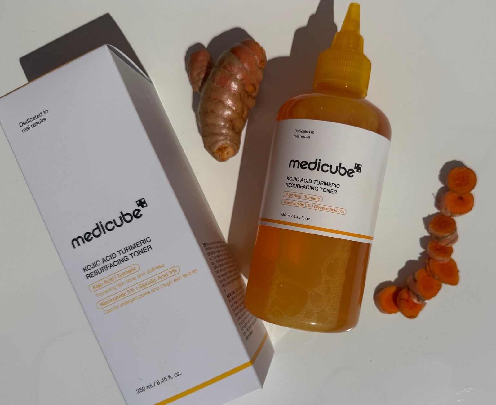 A bottle and packaging box of Medicube Kojic Acid Turmeric Resurfacing Toner next to fresh turmeric root slices