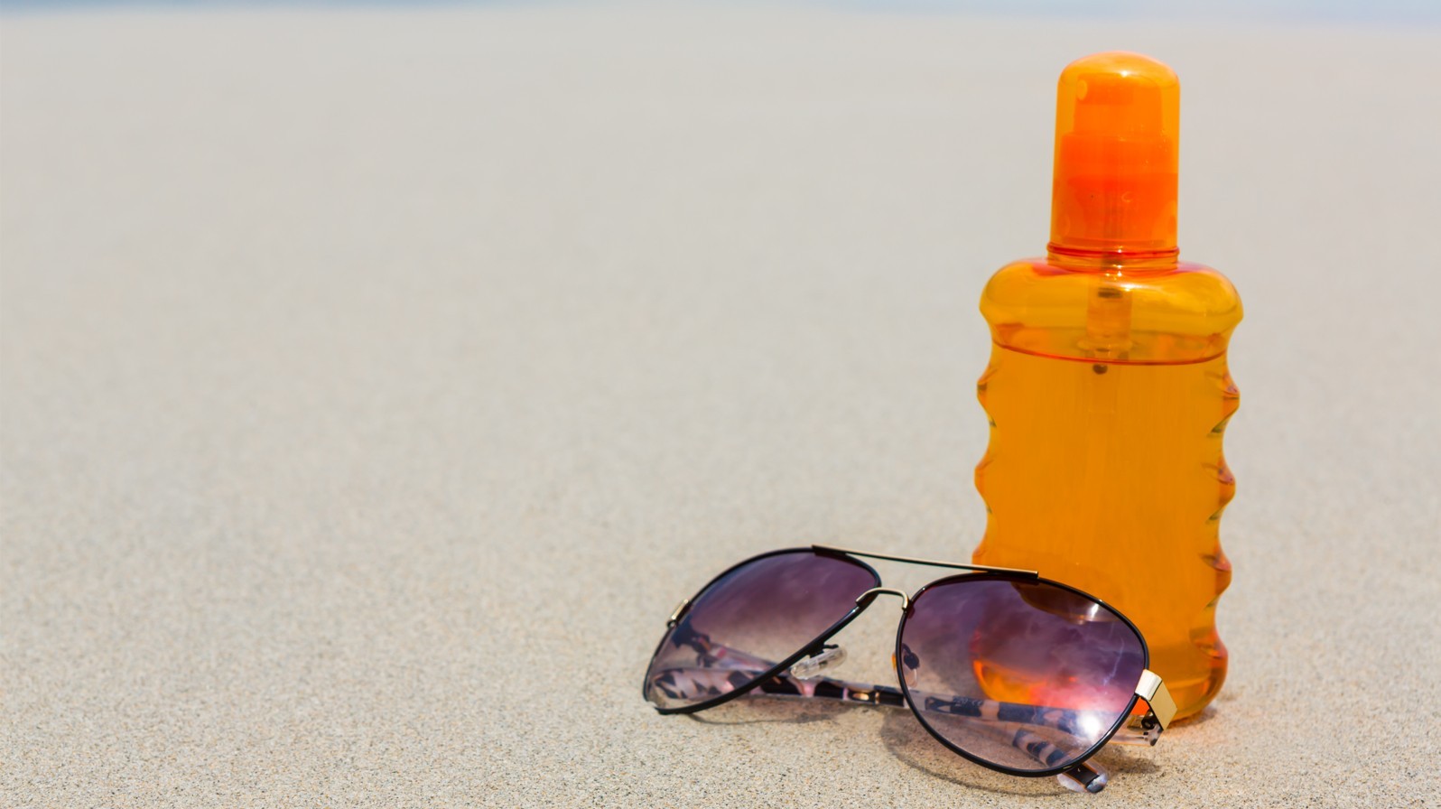 Sunscreen oil bottle sitting on the beach next to purple aviator sunglasses