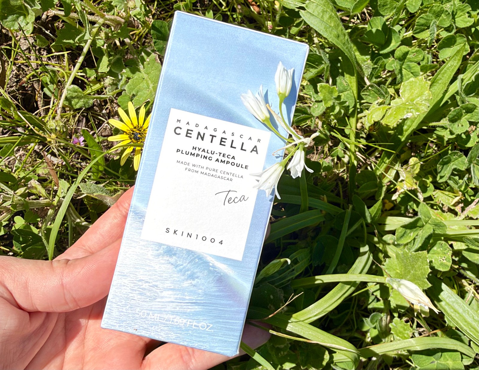 Hand holding SKIN1004 Madagascar Centella Hyalu-Teca Plumping Ampoule box with white flowers outdoors
