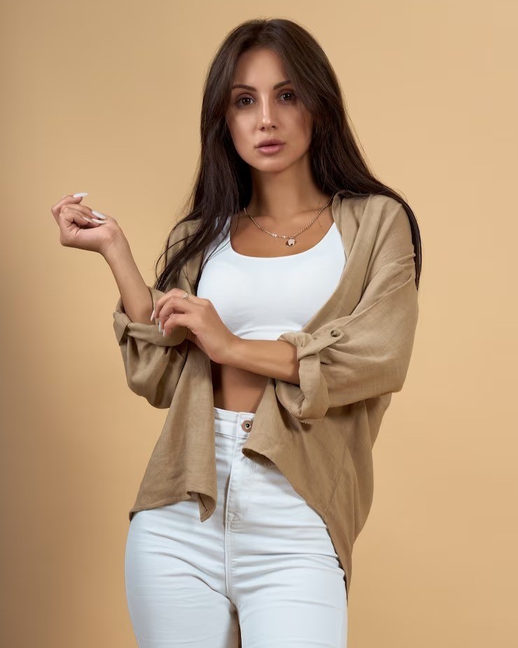 Woman with dark hair posing in front of camera with white singlet and beige overshirt