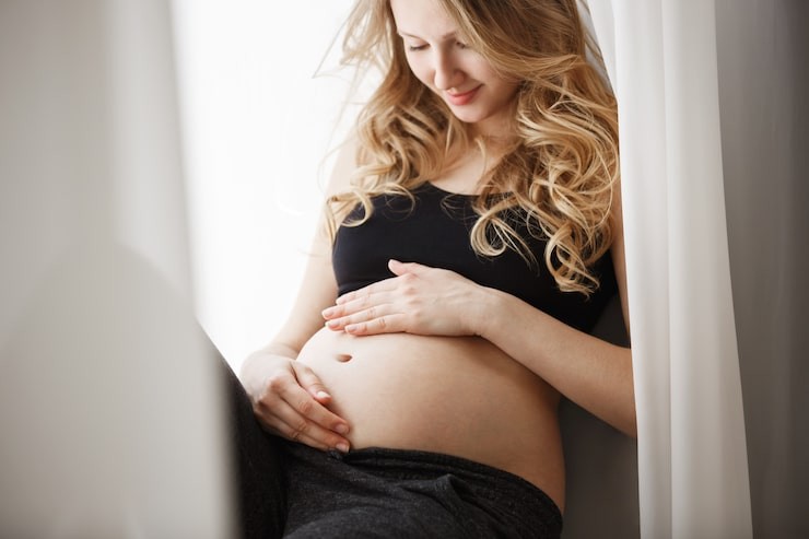 Blonde pregnant woman sitting on a window sill, holding her belly