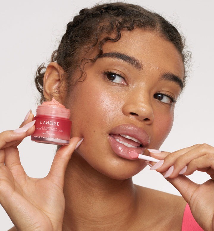 Young model with dark curls holding berry-pink Laneige Lip Sleeping Mask EX in one hand and applying it to her lips with the other