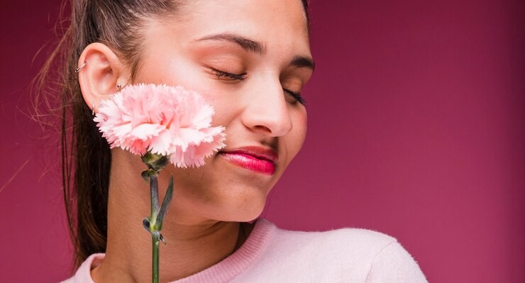 Young woman wearing pink holding a pink carnation to her cheek on a maroon background