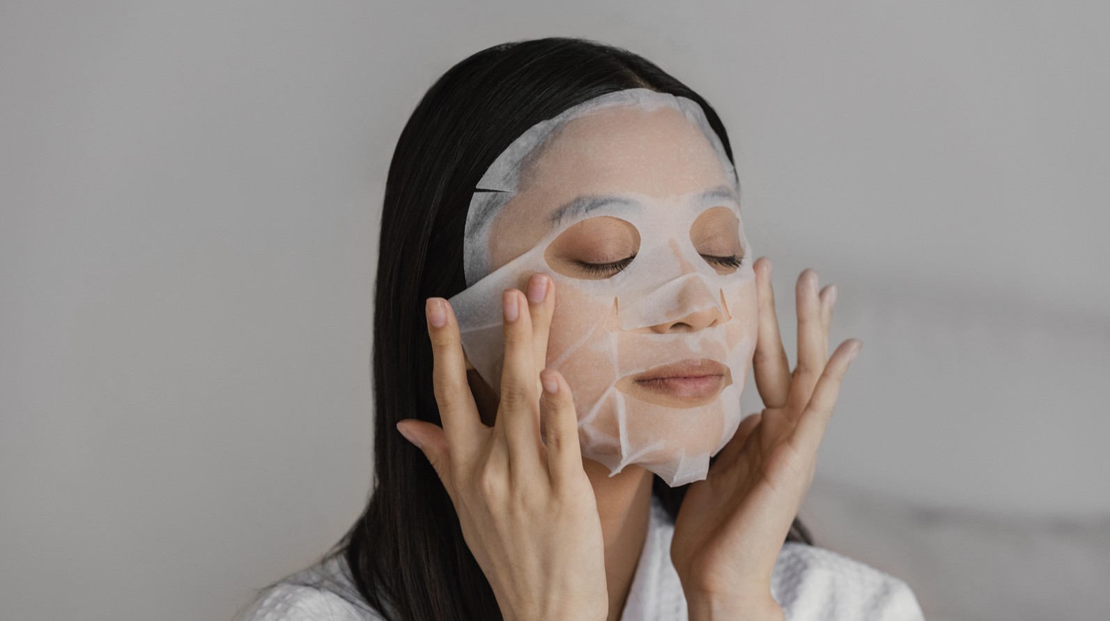Young lady pressing a white sheet mask to her face, eyes closed