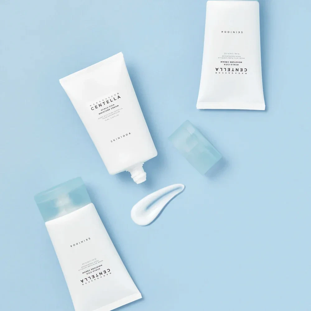 Skin1004 Madagascar Centella Hyalu-Cica Moisture Cream bottles lying on blue floor with test smear of white jelly-like cream
